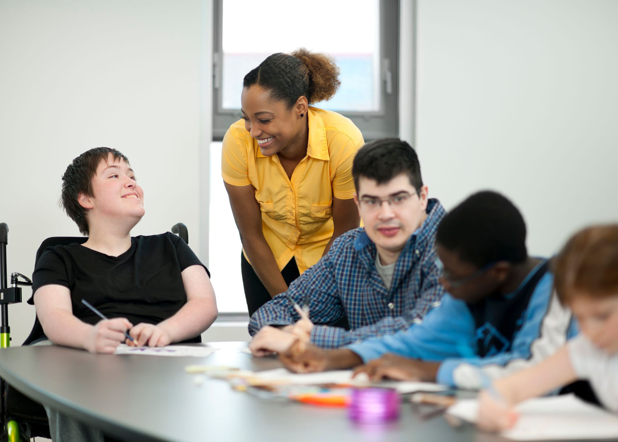 Teacher smiling at a student as they and other young adults work on an assignment at a table.