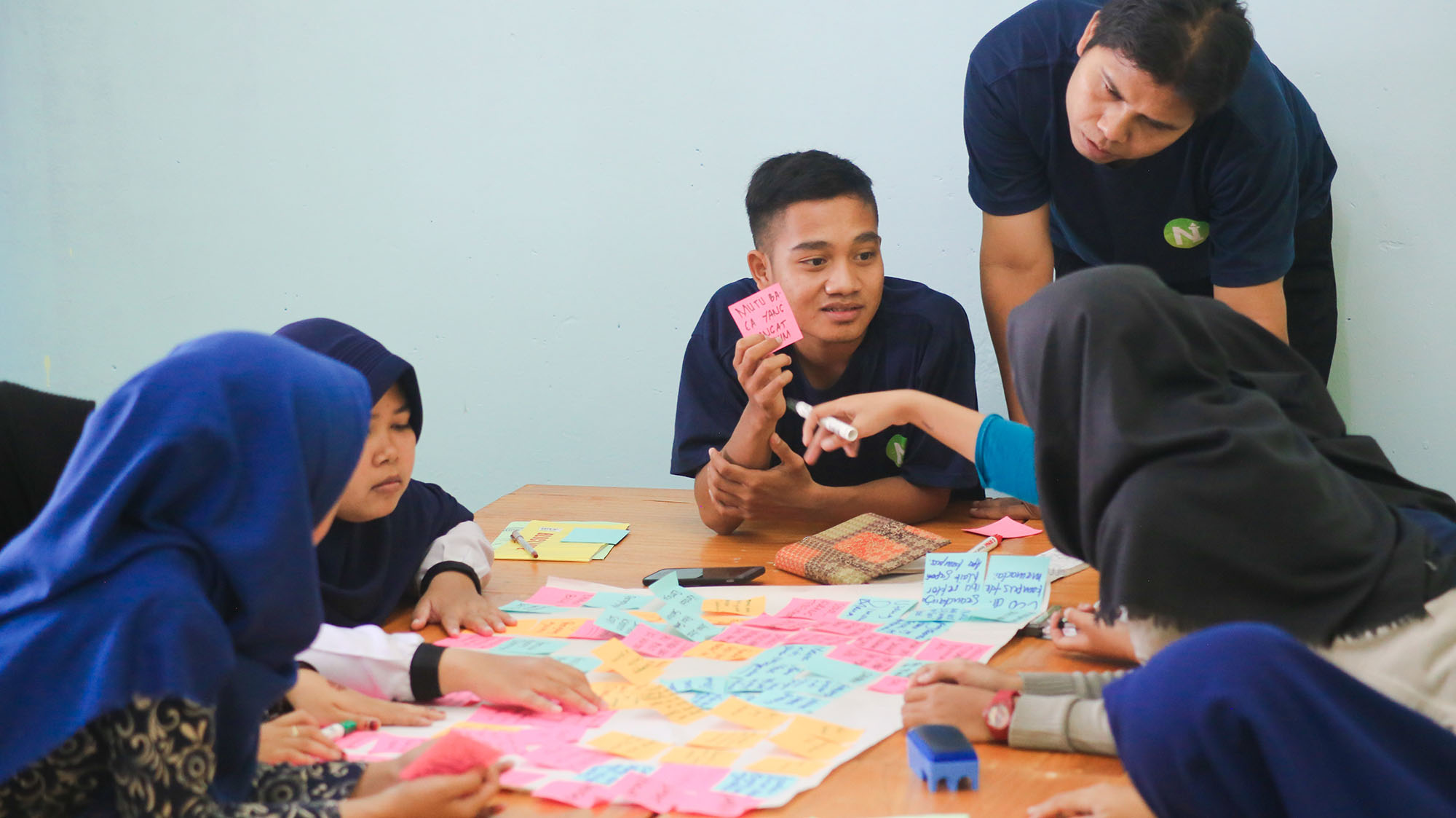 Students gathered around a table, discussing post-its assembled on a large paper.